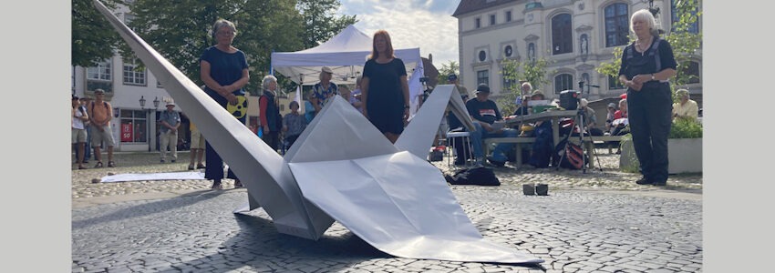   Foto: Dirk Werner. Anlässlich des Gedenktags zu den Atombombenabwürfen über Hiroshima und Nagasaki halten Mitglieder des Friedensbündnisse am 9. August 2025 eine Mahnwache auf dem Marktplatz Lüneburg. Vor ihnen liegt ein gefalteter Kranich aus Papier als Friedenssymbol.